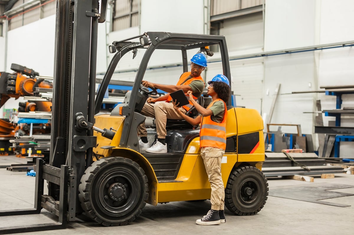 Two multiracial industrial engineers wearing safety vests and helmets are using a digital tablet and driving a forklift truck inside a large factory